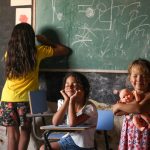 Três crianças participam de uma atividade de aprendizagem diante de um quadro-negro em uma sala de aula.
