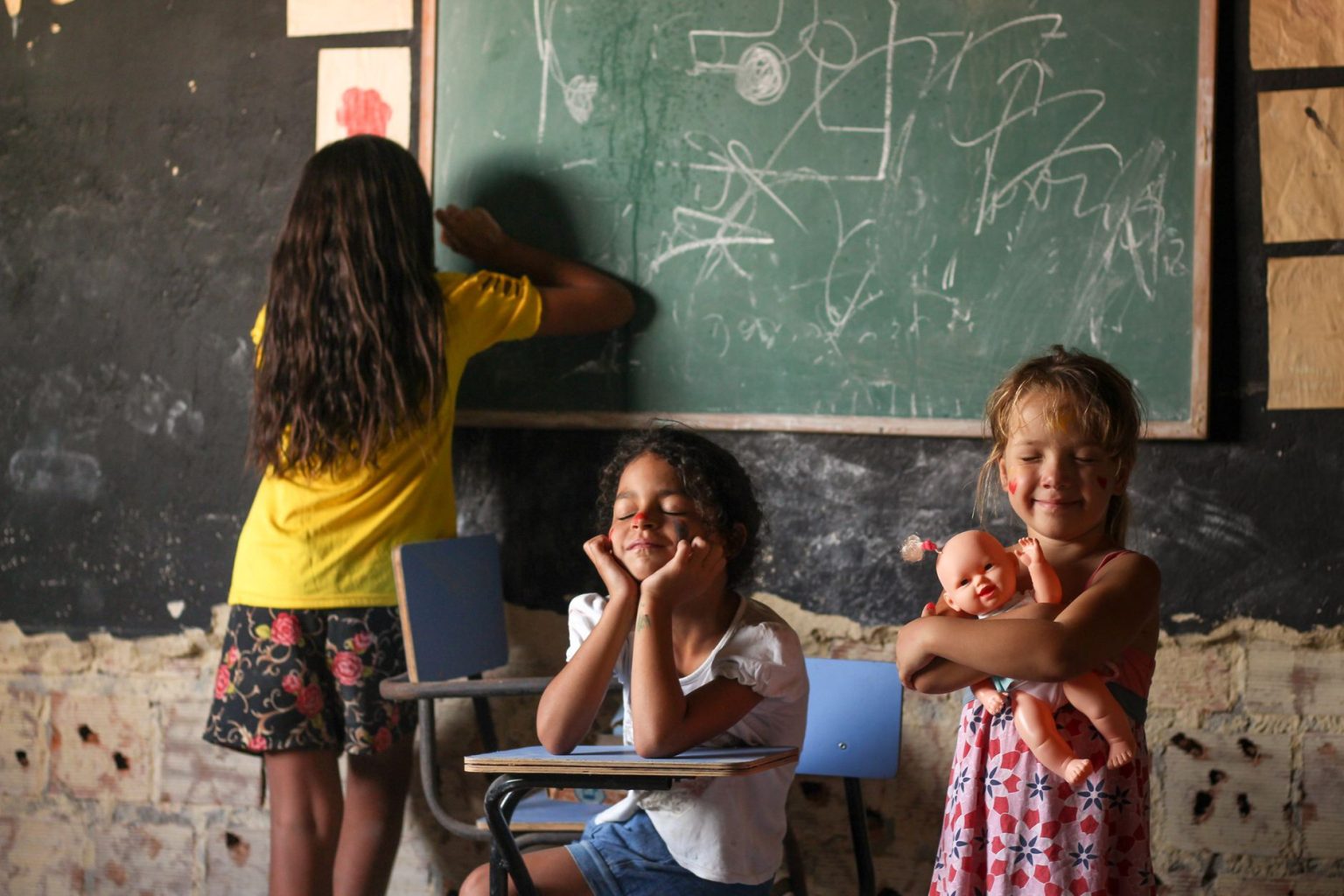 Três crianças participam de uma atividade de aprendizagem diante de um quadro-negro em uma sala de aula.
