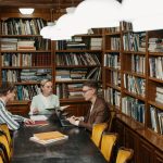 Três profissionais participam de uma reunião com documentos e computadores em uma biblioteca.