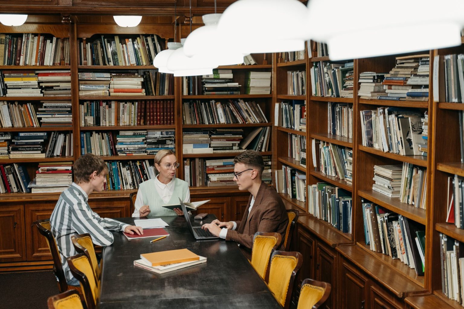 Três profissionais participam de uma reunião com documentos e computadores em uma biblioteca.