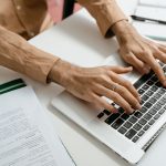 Mãos digitando em um notebook sobre uma mesa com documentos ao redor, representando a adoção de processos administrativos digitais.