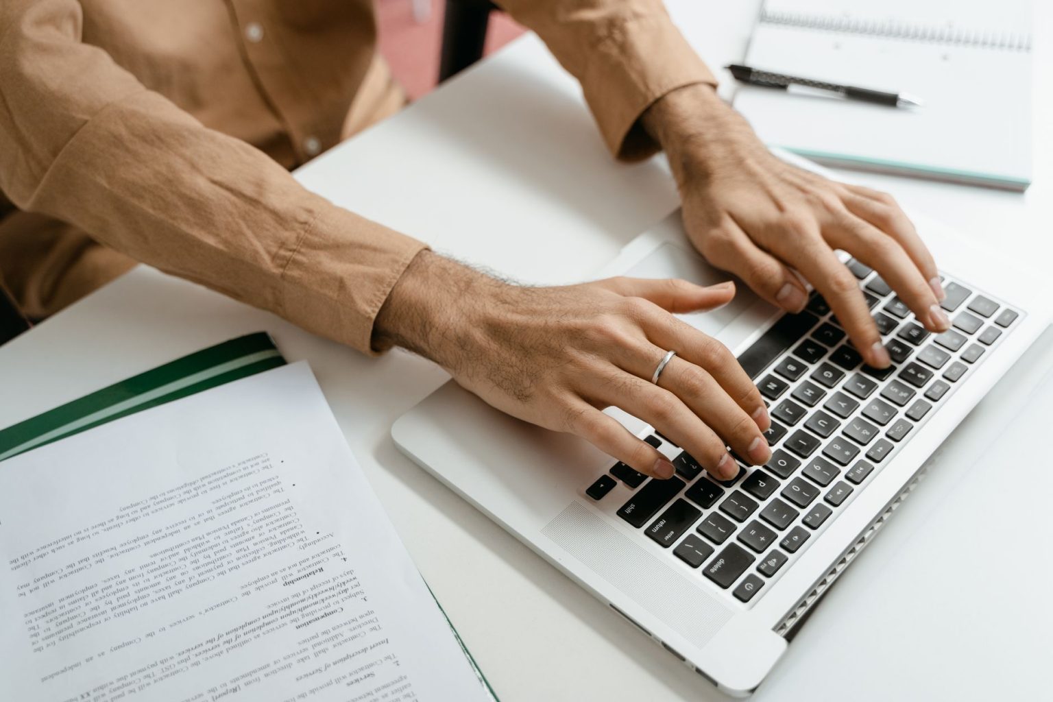 Mãos digitando em um notebook sobre uma mesa com documentos ao redor, representando a adoção de processos administrativos digitais.
