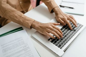Mãos digitando em um notebook sobre uma mesa com documentos ao redor, representando a adoção de processos administrativos digitais.