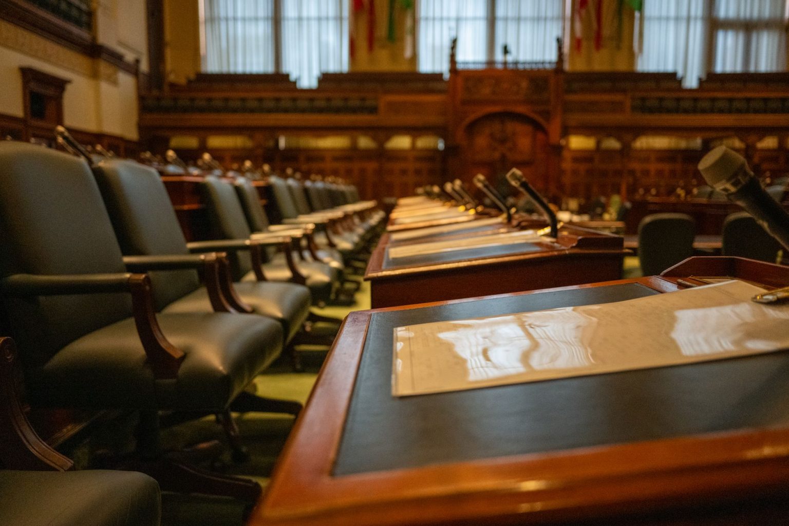 Interior de uma câmara legislativa vazia, com fileiras de mesas e microfones.