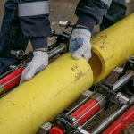 Trabalhador instala tubulações amarelas ao ar livre usando ferramentas e equipamentos.