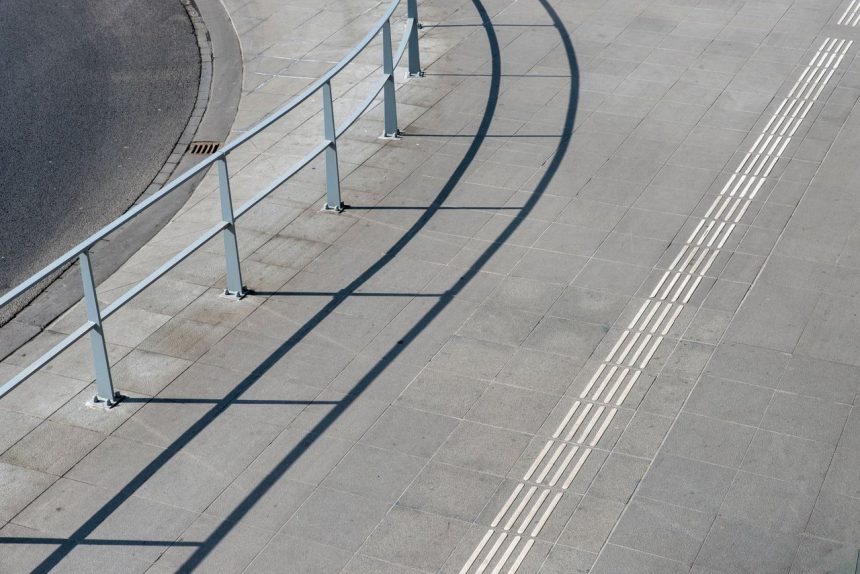 Vista aérea de uma calçada urbana com corrimãos metálicos e piso tátil para acessibilidade.