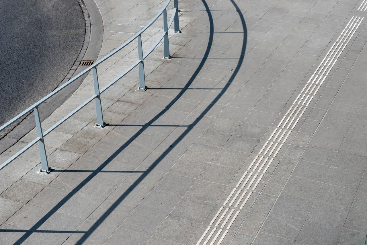 Vista aérea de uma calçada urbana com corrimãos metálicos e piso tátil para acessibilidade.