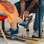 Terapeuta auxilia uma mulher durante sessão de reabilitação com pesos nos tornozelos.